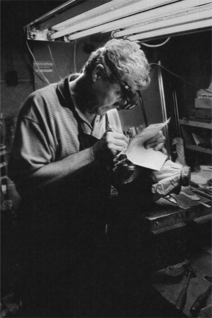 Photographie d'archives en noir et blanc du père de Roland Baptiste, concentré sur un minutieux travail de gravure à son établi, sous la lumière de l'atelier.