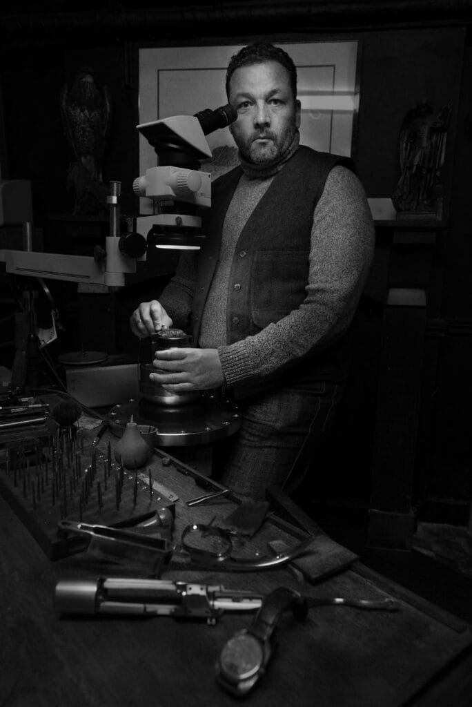 Portrait en noir et blanc de Roland Baptiste, créateur de la marque horlogère indépendante RBaptiste, debout dans son atelier en Belgique. Il tient un appareil photo entre ses mains, entouré de ses outils de précision.