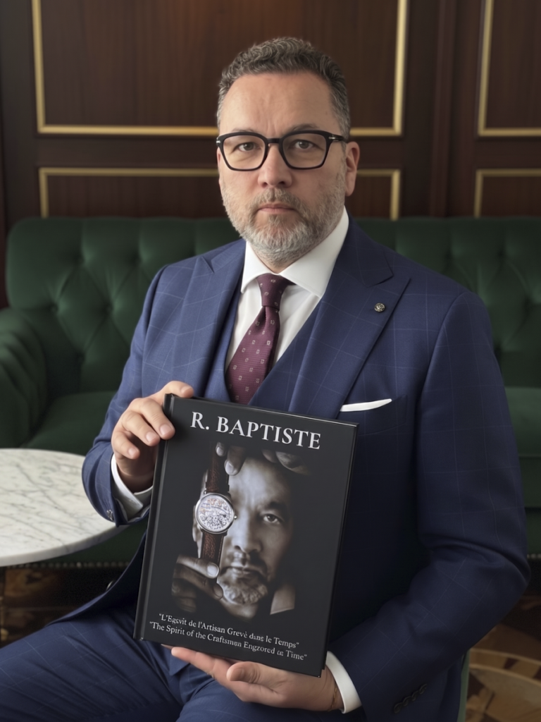 Portrait de Roland Baptiste en costume trois pièces bleu, assis et présentant la couverture de son livre d'art intitulé "R. BAPTISTE". Sur la couverture du livre, on aperçoit un portrait en noir et blanc d'un artisan tenant une montre gravée. En arrière-plan, un canapé en velours vert et des boiseries.