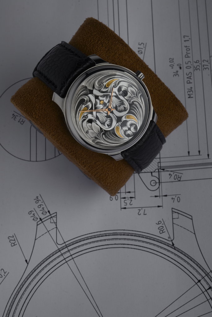 Montre RBaptiste posée sur des plans techniques de conception horlogère. Cadran avec gravure profonde à la main et détails en or, réalisée par l'horloger créateur Roland Baptiste.