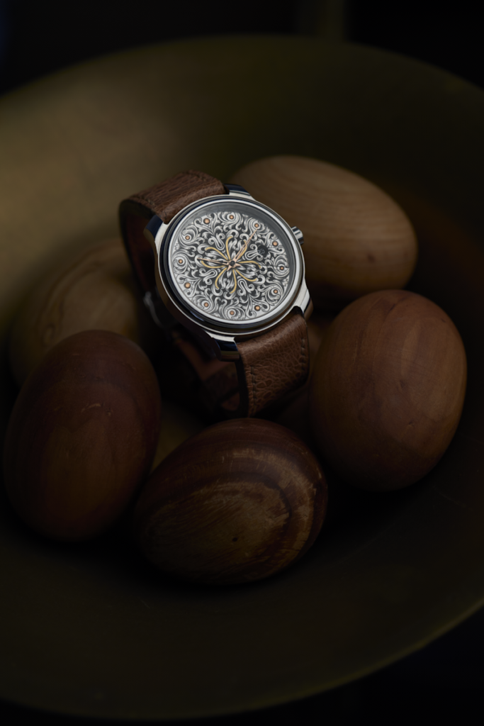 Montre RBaptiste par Roland Baptiste, horloger créateur indépendant. Cadran minutieusement gravé à la main au burin avec des accents dorés, posée au milieu d'éléments naturels en bois poli.