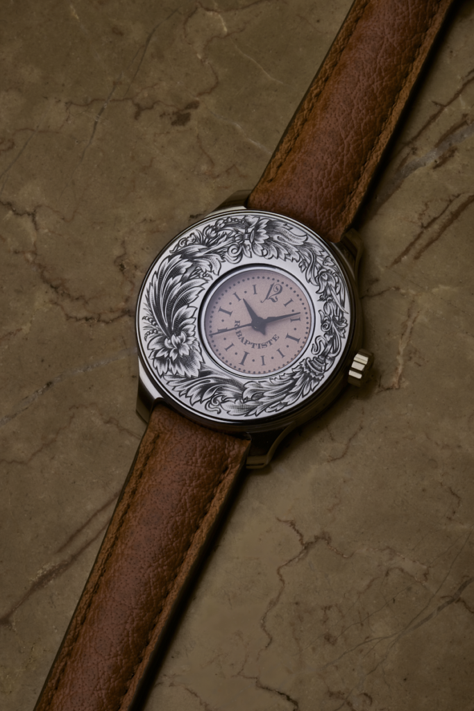 Montre Audace 34 mm avec cadran excentré rose poudré et boîtier gravé de volutes, posée sur un sol aride et texturé.