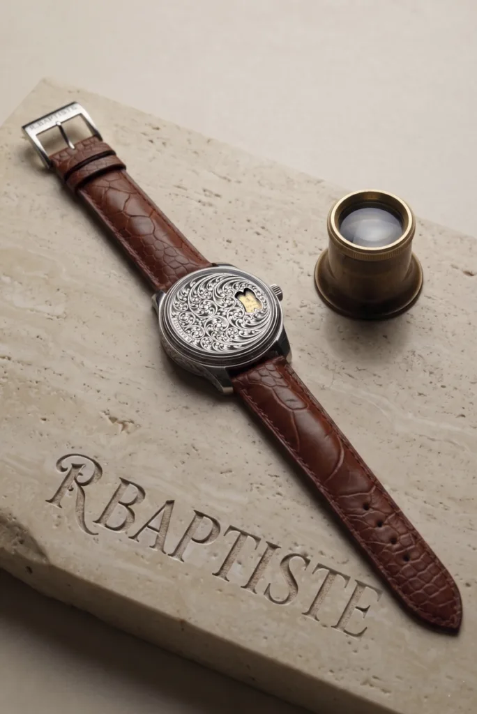 Gros plan sur une montre de luxe RBaptiste. Le cadran est une œuvre d'art complexe, symétriquement gravée à la main au burin avec des motifs ornementaux et des aiguilles dorées. Boîtier en acier poli et bracelet noir texturé. Sur un fond d'écrin de marque.